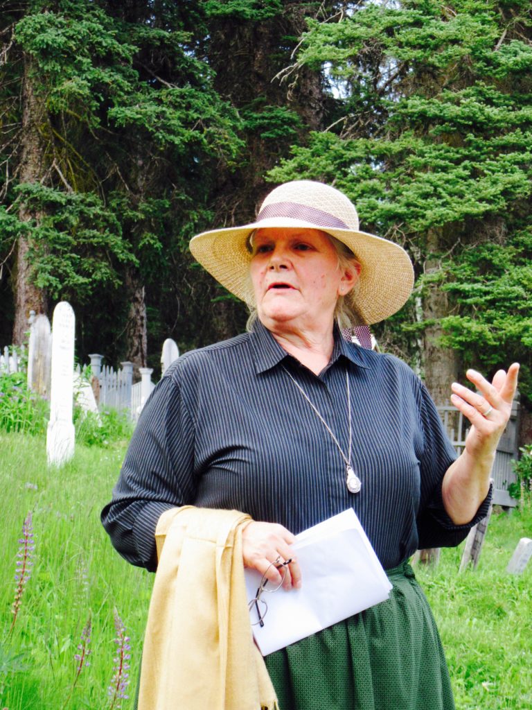 Woman in large straw hat, dark grey shirt and long green skirt holding a paper and standing in front of tombstones.