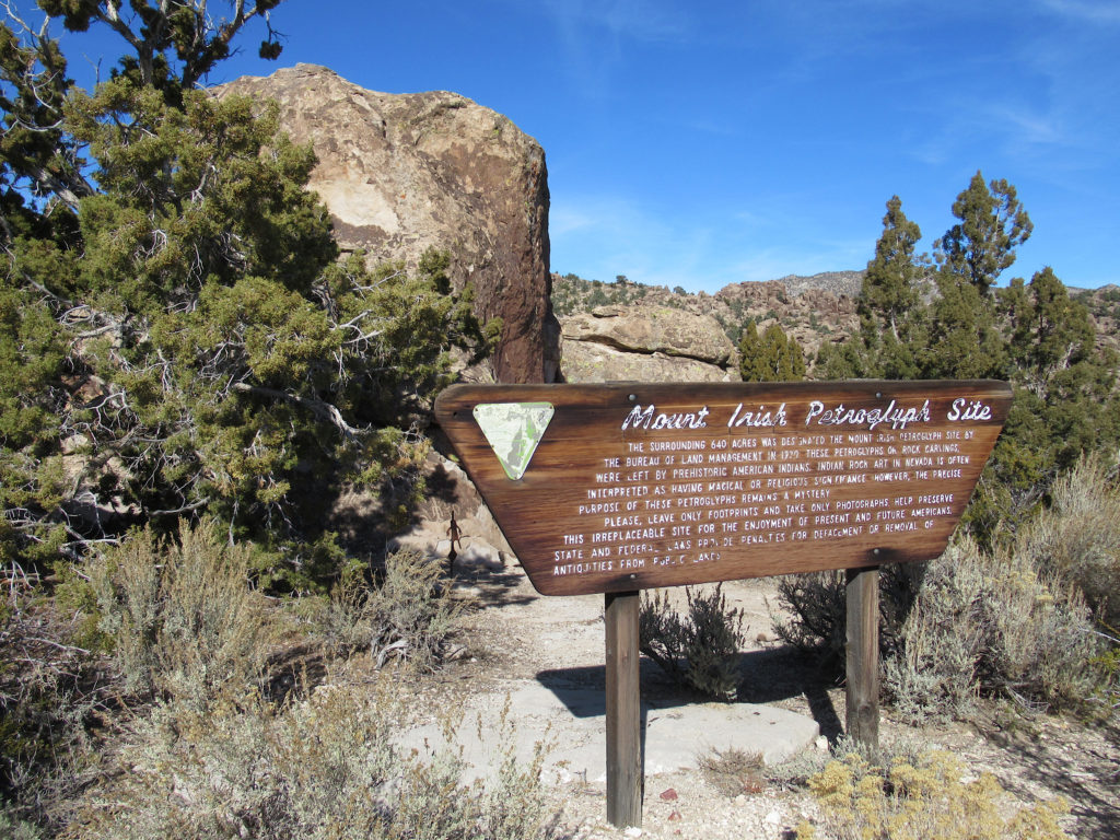 A wooden sign, reading Mount Irish Petroglyph Site, stands in front of a large rock with juniper trees all around it
