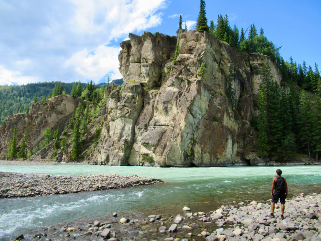 Man standing beside confluence of two light blue rivers with large cliff in background.