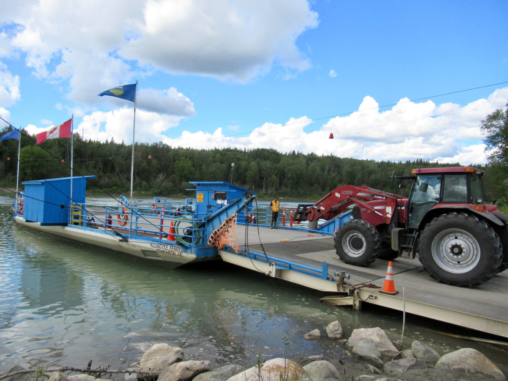 Red tractor driving on to small blue ferry with Canadian and Albertan flags.