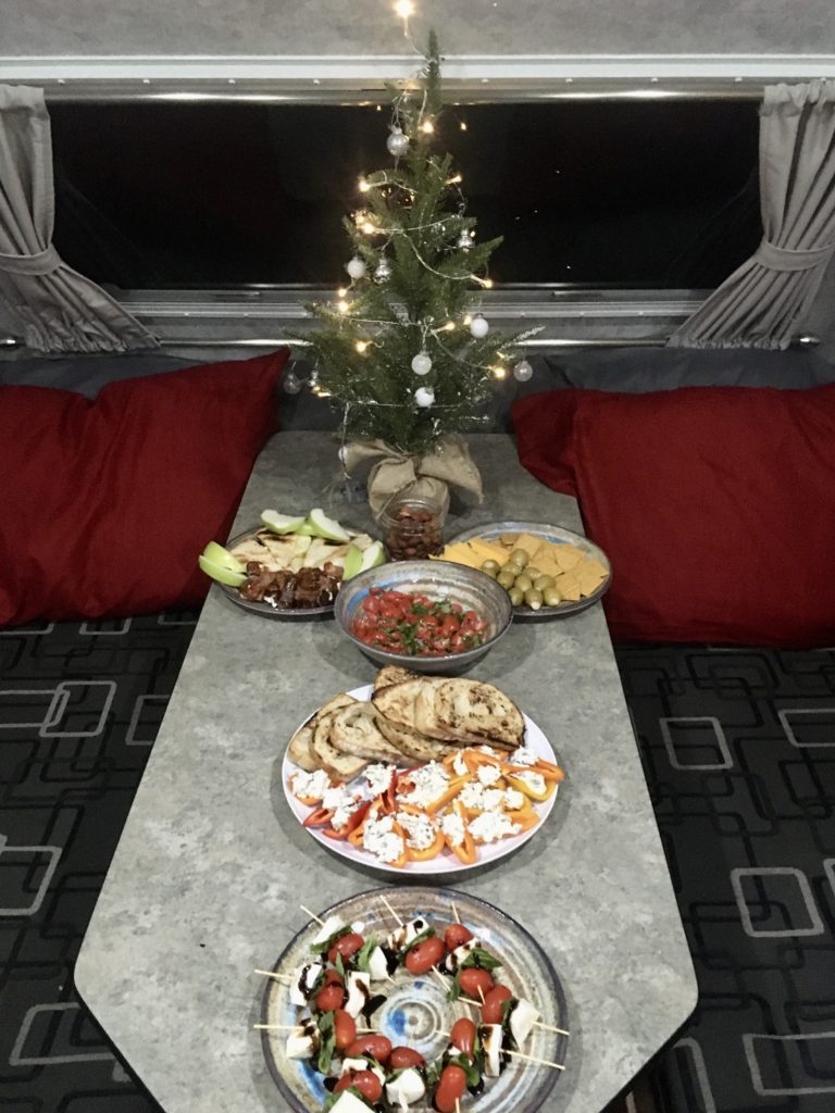 Trailer table loaded with plates of food and a small, artificial Christmas tree. Red pillows on grey seat cushions in background.