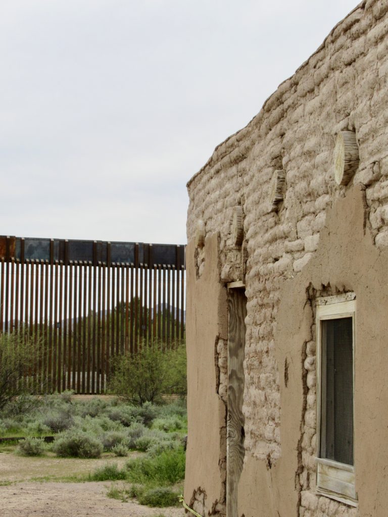 Brown adobe building in foreground with the metal border wall in background.