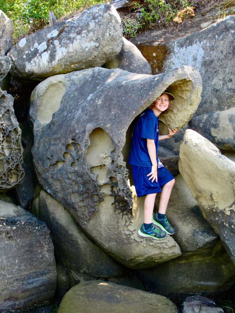 Young boy in blue shirt and shorts standing in mouth of rock that looks like a fish.