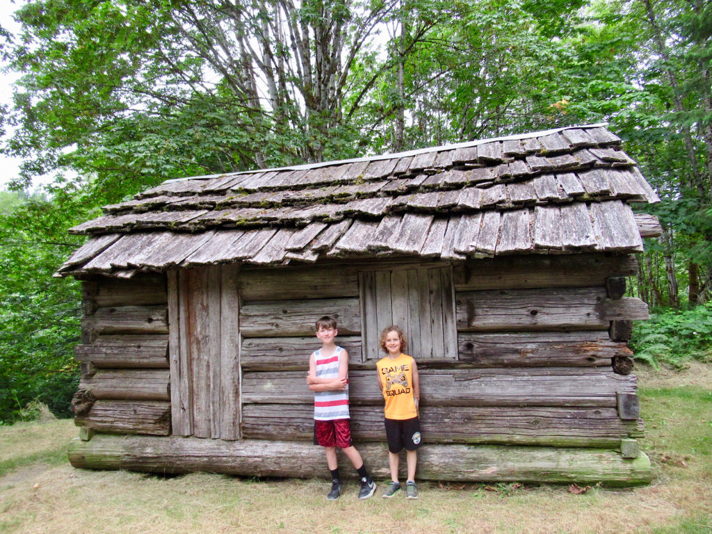 Two young boys in shorts and t-shirts standing beside historic wooden cabin.