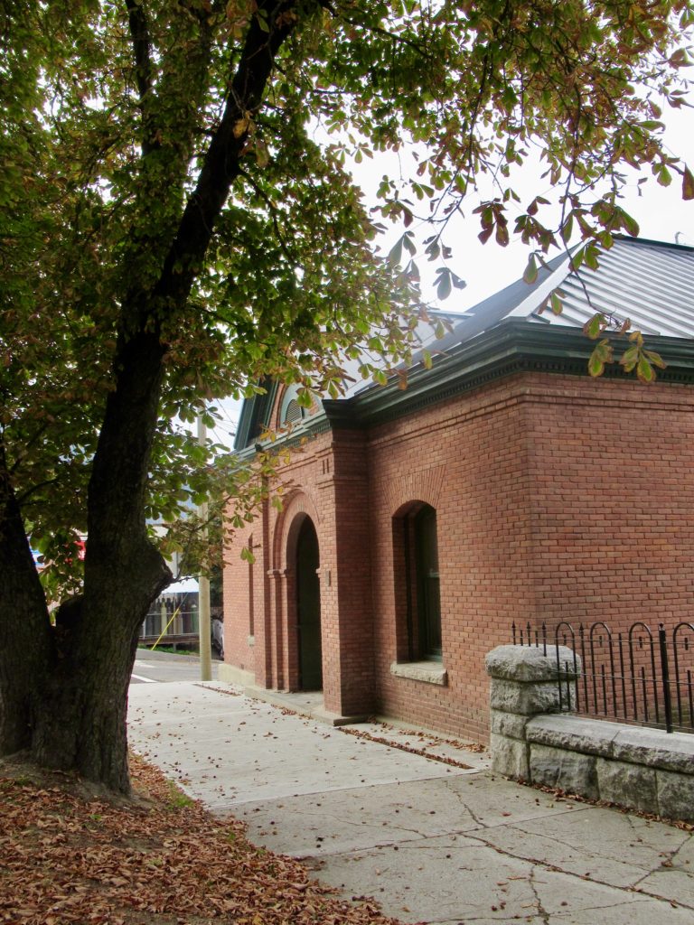 Brick building with green metal roof off sidewalk with large tree on lefthand side of photo.