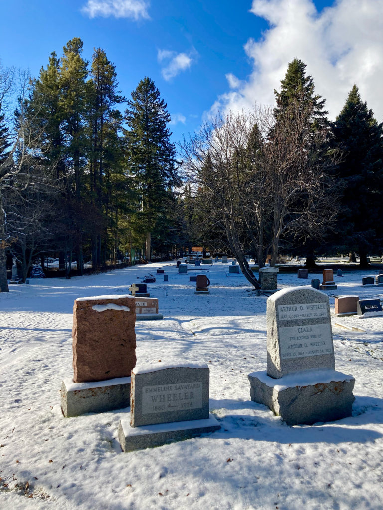 Three large headstones in a snow-covered cemetery under a bright blue sky.