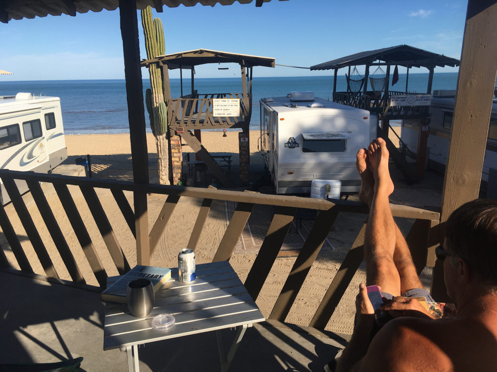 Man sitting on a wooden deck with feet up on railing overlooking trailers, sand and the ocean.