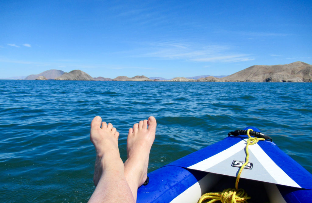 Bare feet propped up on blue inflatable kayak with ocean and distant islands in background.
