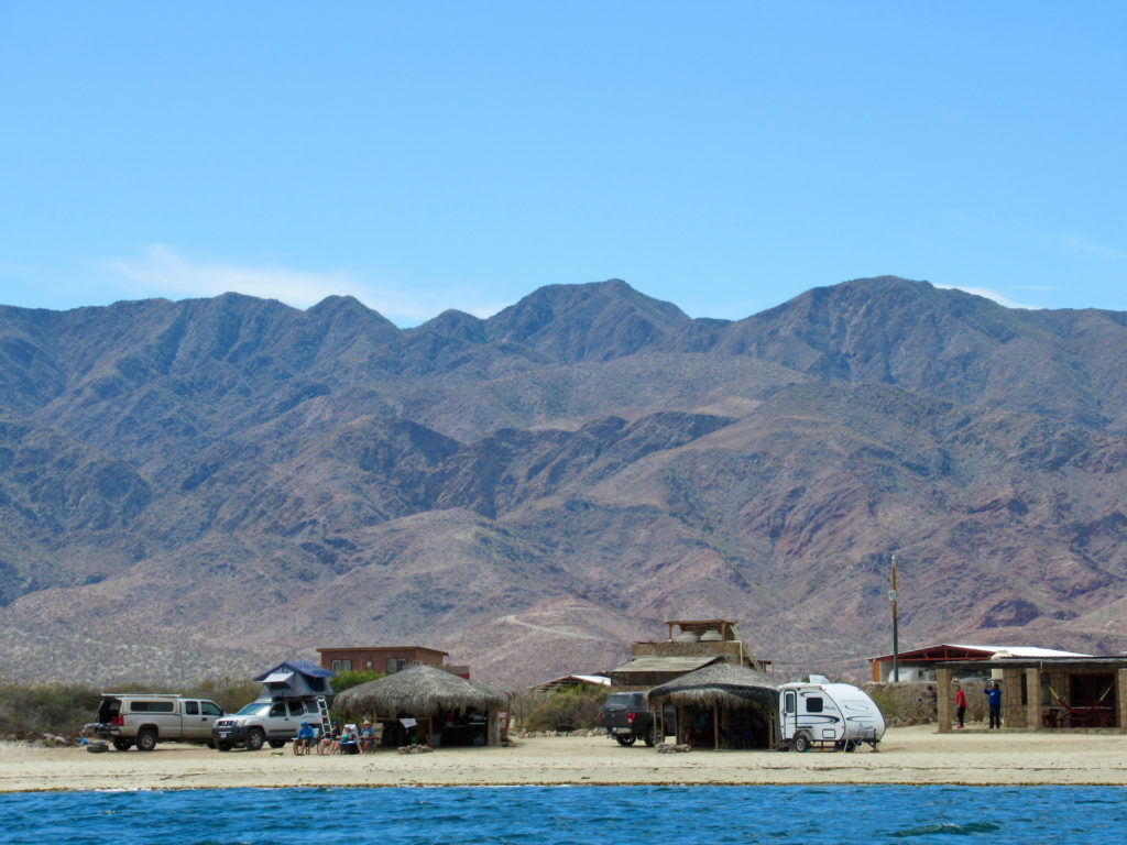 Trailers and other camping units on beach with palapas. Tall mountains in background and ocean in foreground.