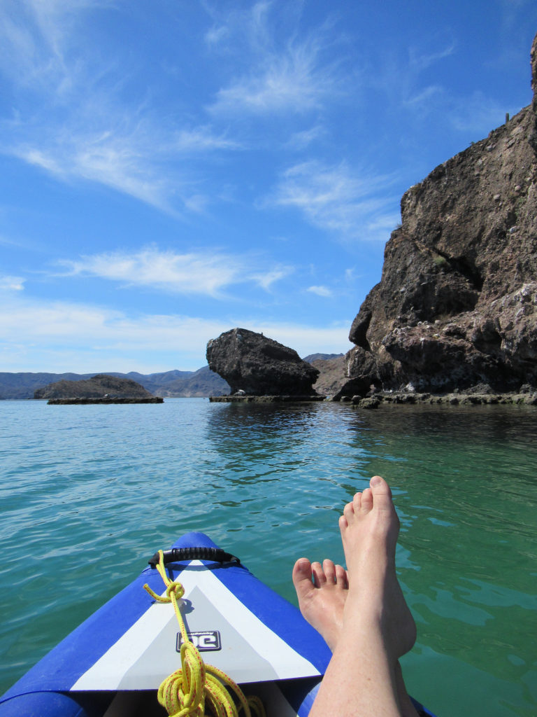 Two crossed feet on bow of blue inflatable kayak on blue green waters with dark lava island in background.