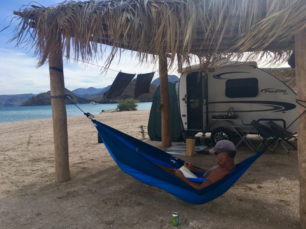 Man lying in blue hammock and reading a book under a palm palapa with small trailer in background.