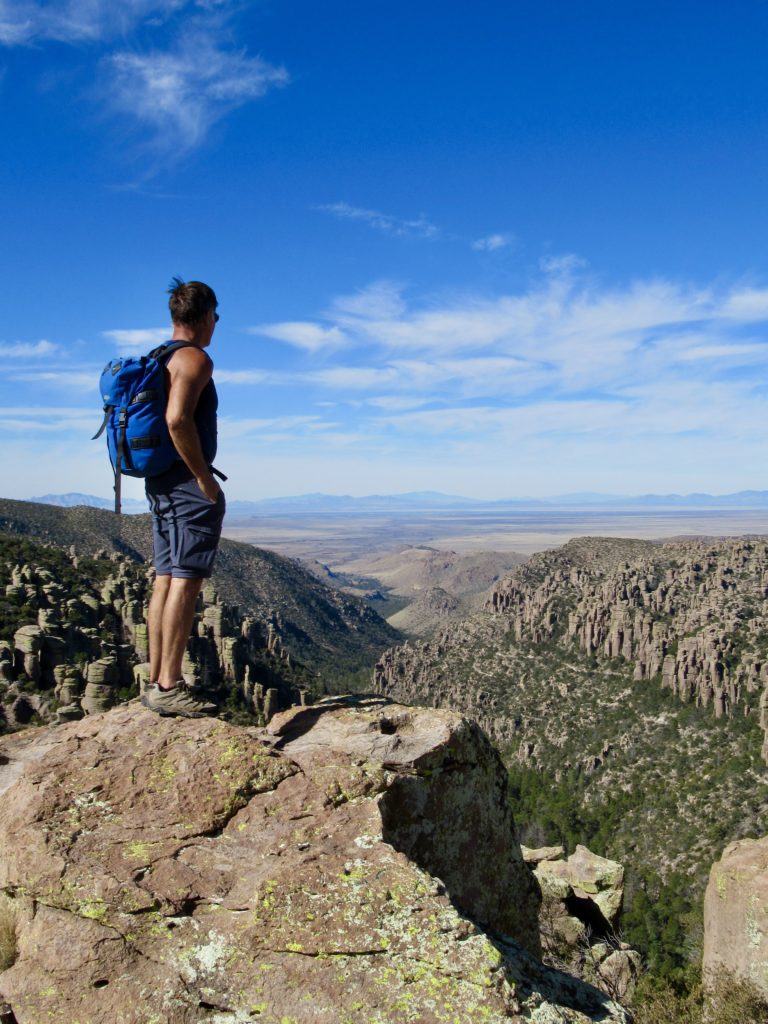Man in blue short and tank top with blue backpack standing on a rocky overlook.