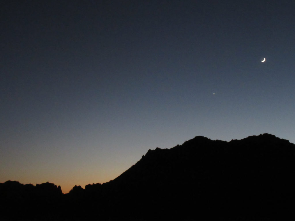 Black silhouette of mountain with moon and planet overhead in evening sky touch by last glow of sunset.