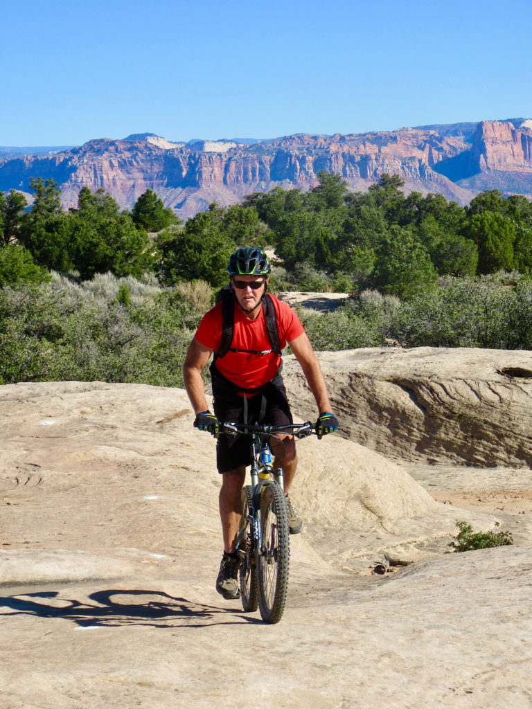 Man in red shirt and black short with green bicycle helmet riding over slick rock with red cliffs in distance.