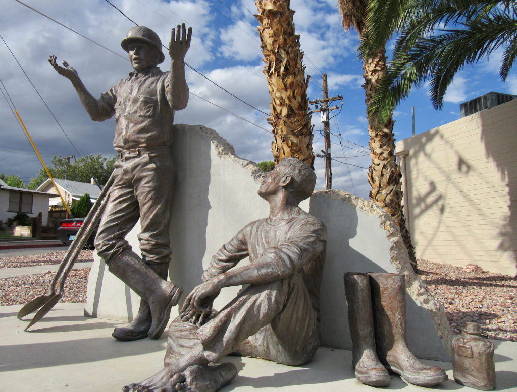 Bronze statue of two workers, one standing with two shovels and wearing a hardhat and the other sitting crosslegged with boots off and a water canteen beside him.