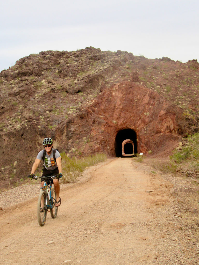 Man on bicycle riding towards photographer with abandoned railway tunnels carved in rock behind him.