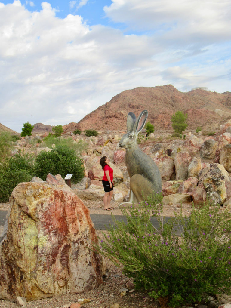 Women in red shirt and black shorts standing face to face with 10-foot high jackrabbit sculpture in red rock boulder garden.