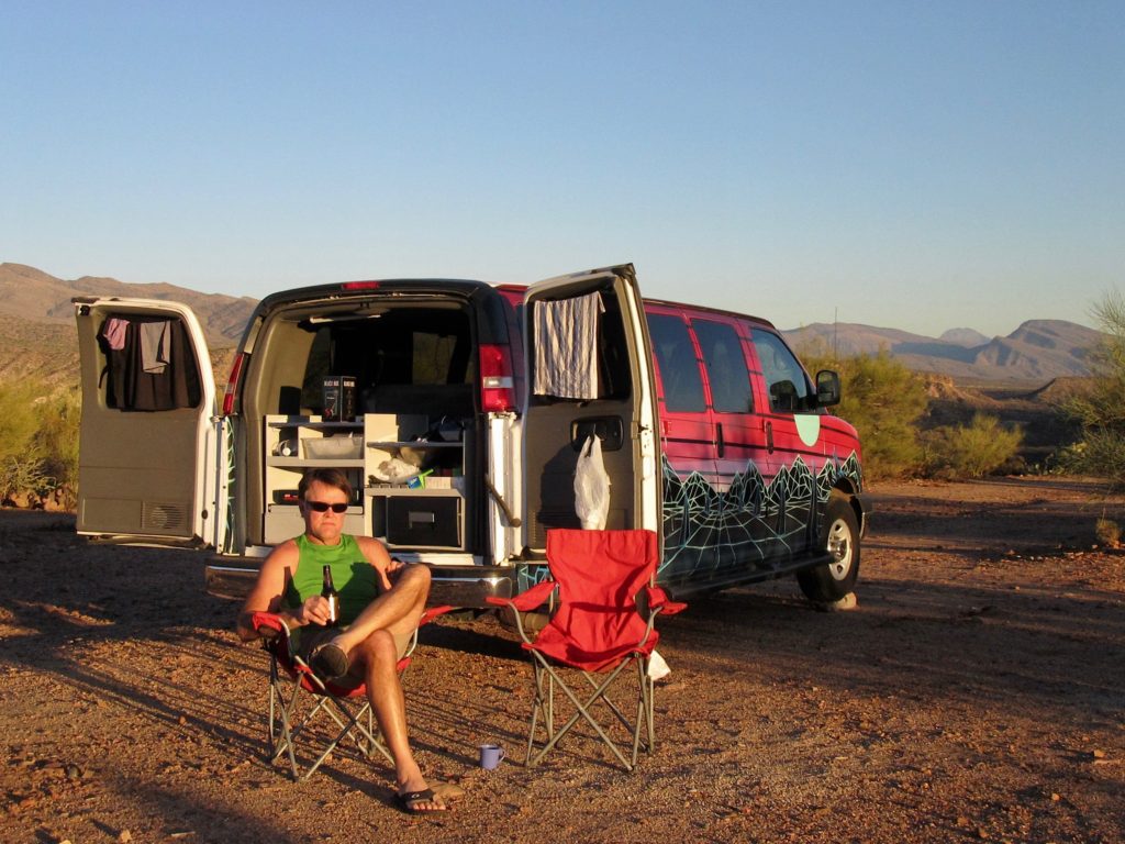 Man sitting in red camp chair with a beer in front of camper van with back doors open.