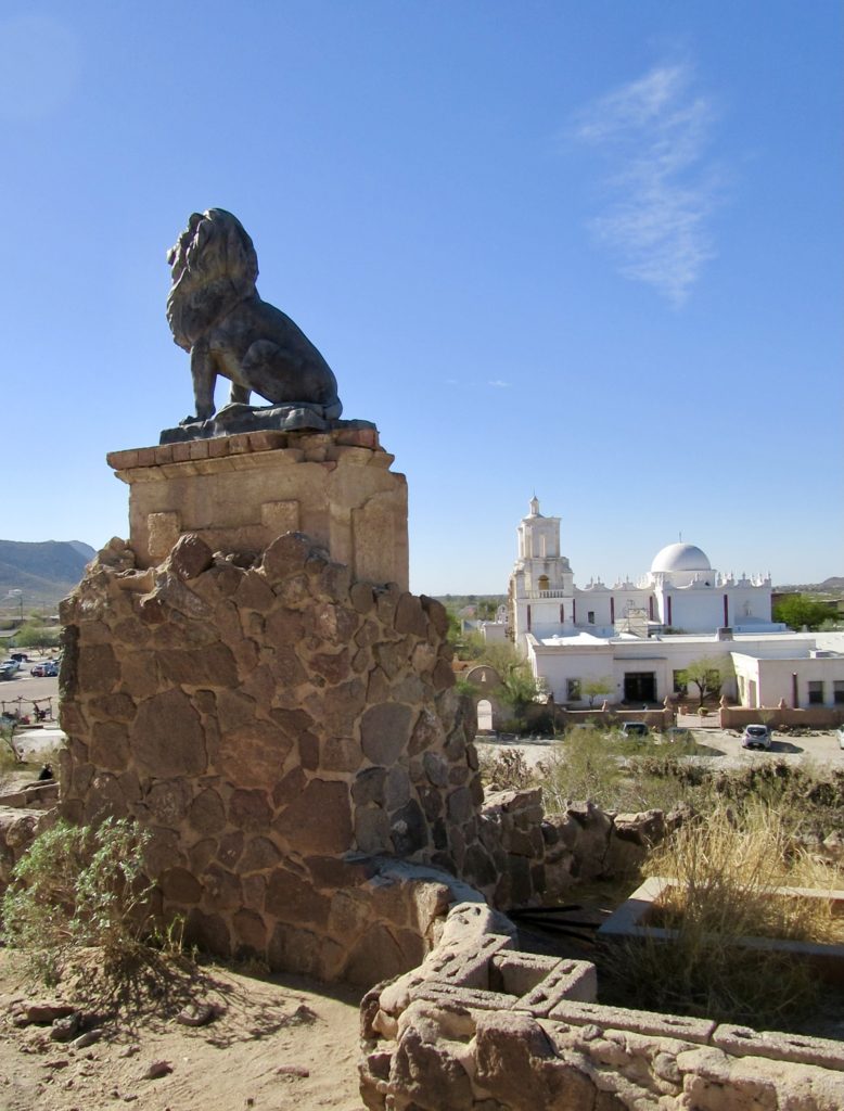 Lion statue on pedestal overlooking white washed mission building beneath blue sky.