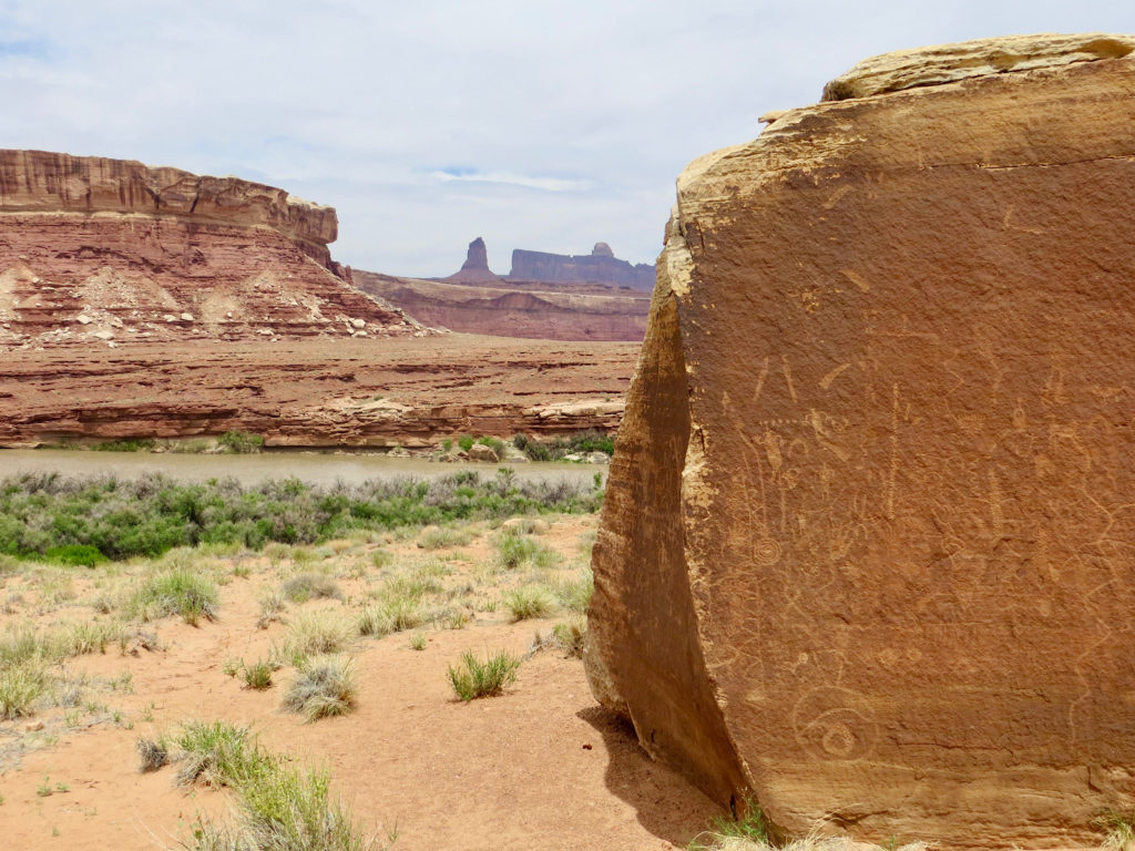 Large sandstone boulder with inscribed shapes and figures on sandy slope above river and with red hills in distance.