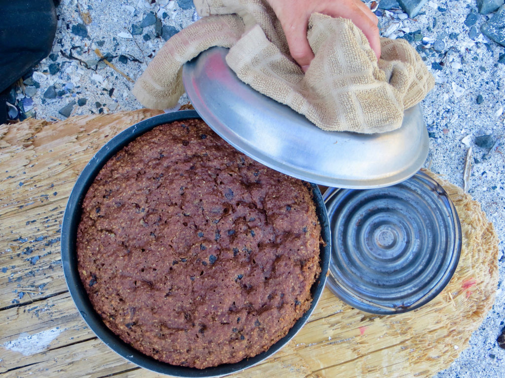 Cake baked in a stovetop oven with woman's hand holding lid and small metal heat diffuser off to the side. 