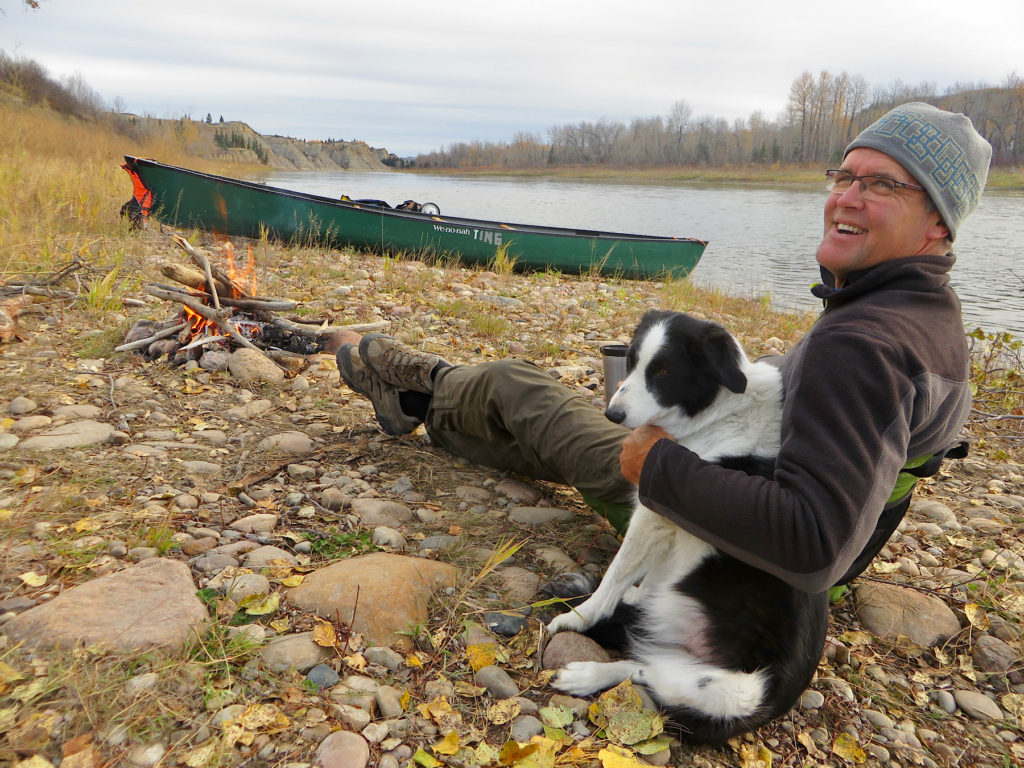 Man and border collie dog sitting in front of a campfire with green canoe in distance pulled up from river.