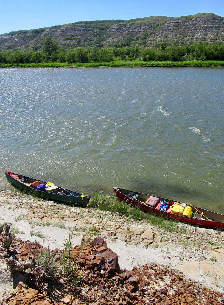 Loaded green and red canoes tied up on shore of wide river with red rocks in foreground. 