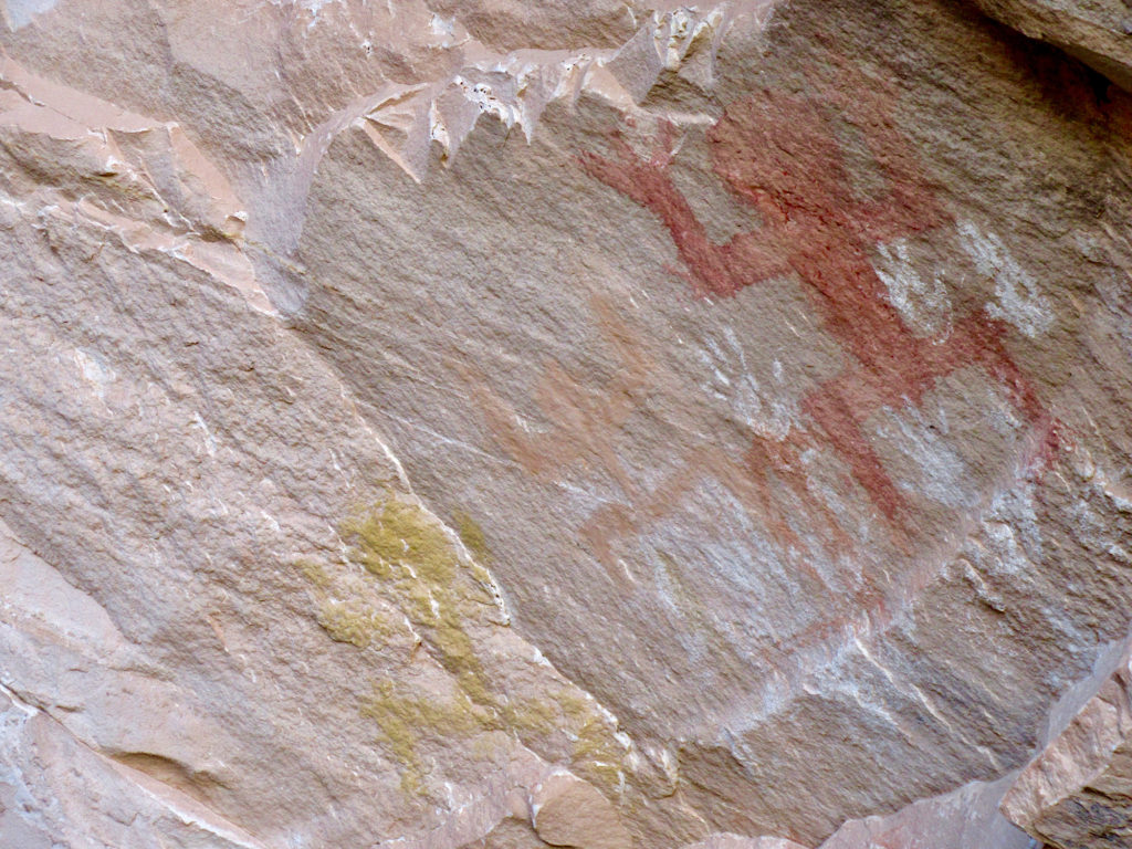 Red, orange and yellowish images of human-like figures painted on rock wall.