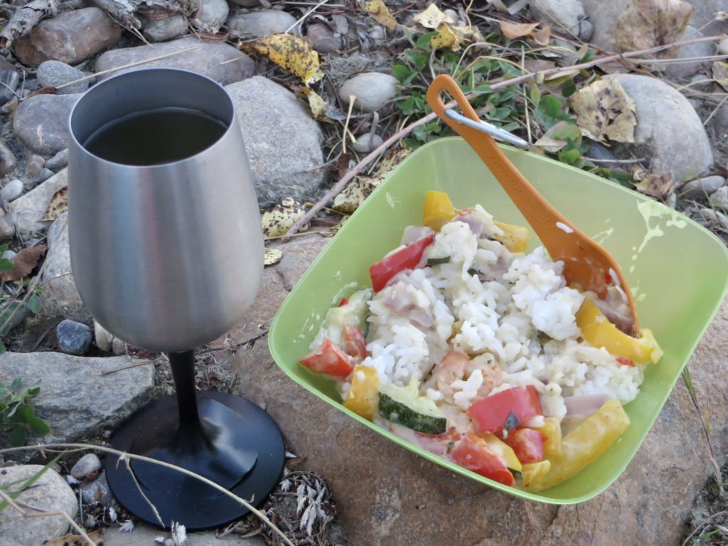Metal wine glass beside green plastic bowl with orange spoon and rice dish with prawns and red and yellow peppers.