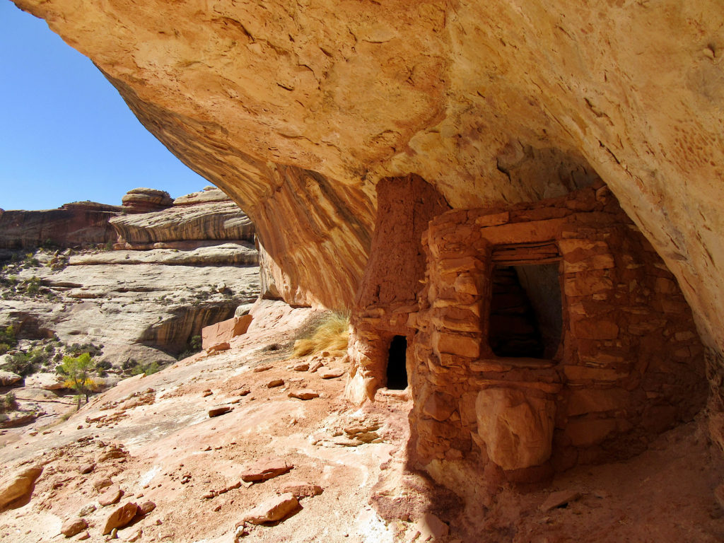 Ancient red rock buildings tucked under overhanging buff-coloured sandstone wall in a canyon.