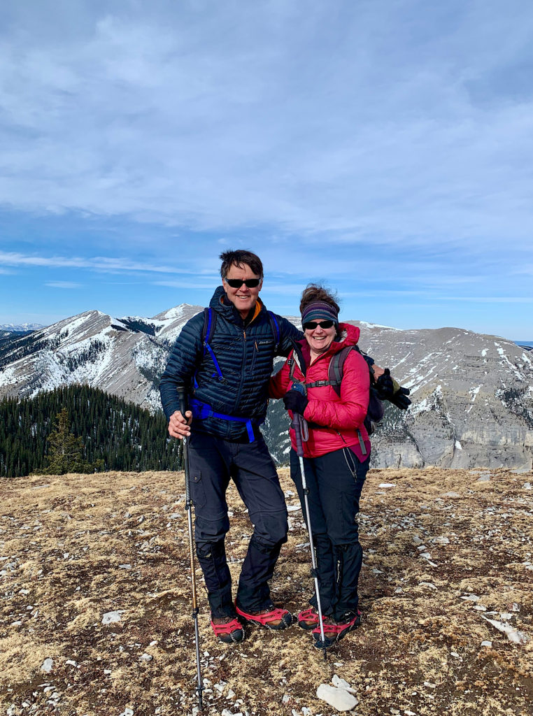 Man in dark jacket and woman in red jacket standing on grassy mountaintop holding hiking poles.