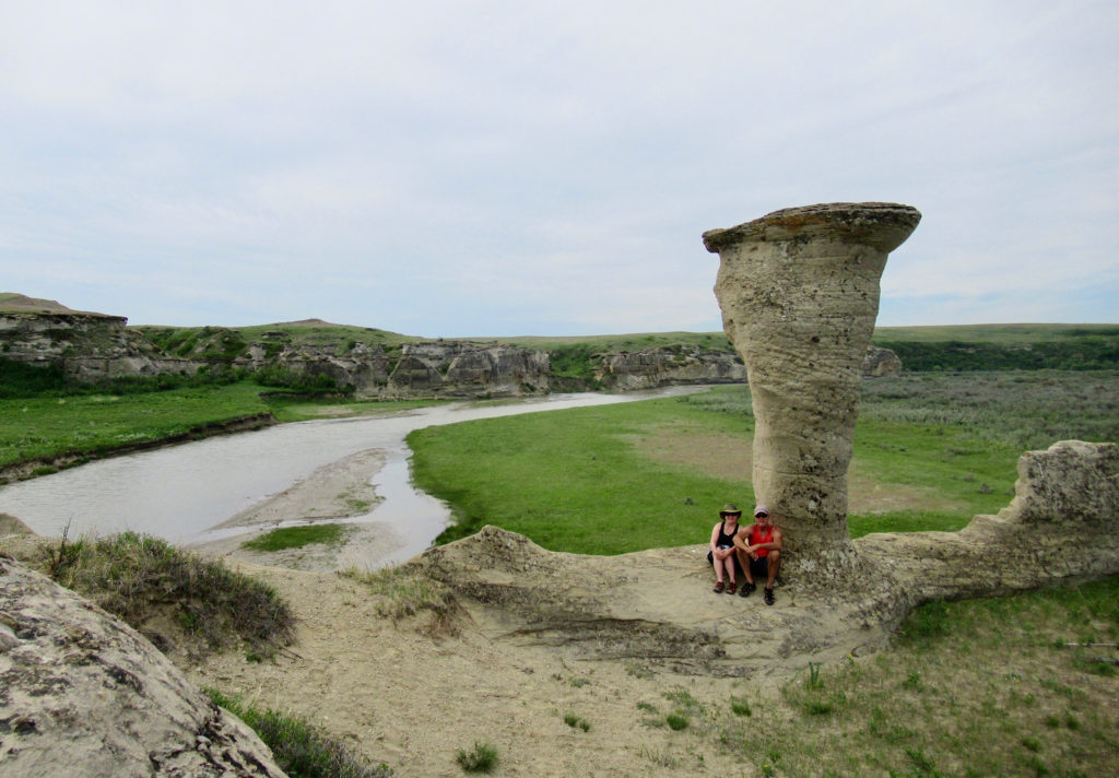 Two people in shorts sitting at base of tall beige sandstone hoodoo above river.