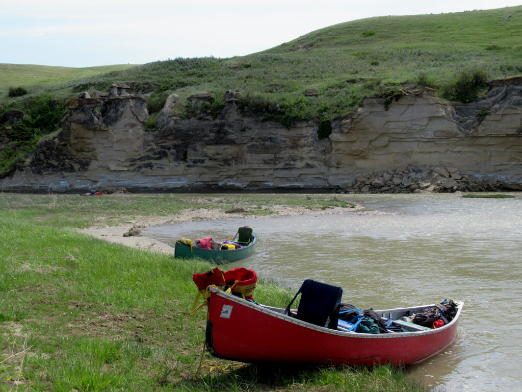 Red and green canoes loaded with gear and pulled up on shore of milky brown river.