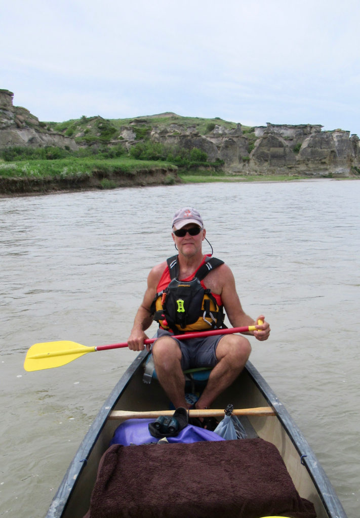 Man in canoe wearing personal floatation device over shorts and short-sleeved top sitting with yellow paddle resting on canoe gunnel.