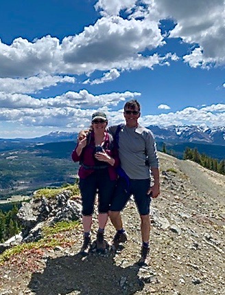 Man and woman in shorts and long sleeved shirts with hats and sunglasses standing on rock outcrop high above evergreen forest with mountains in background.