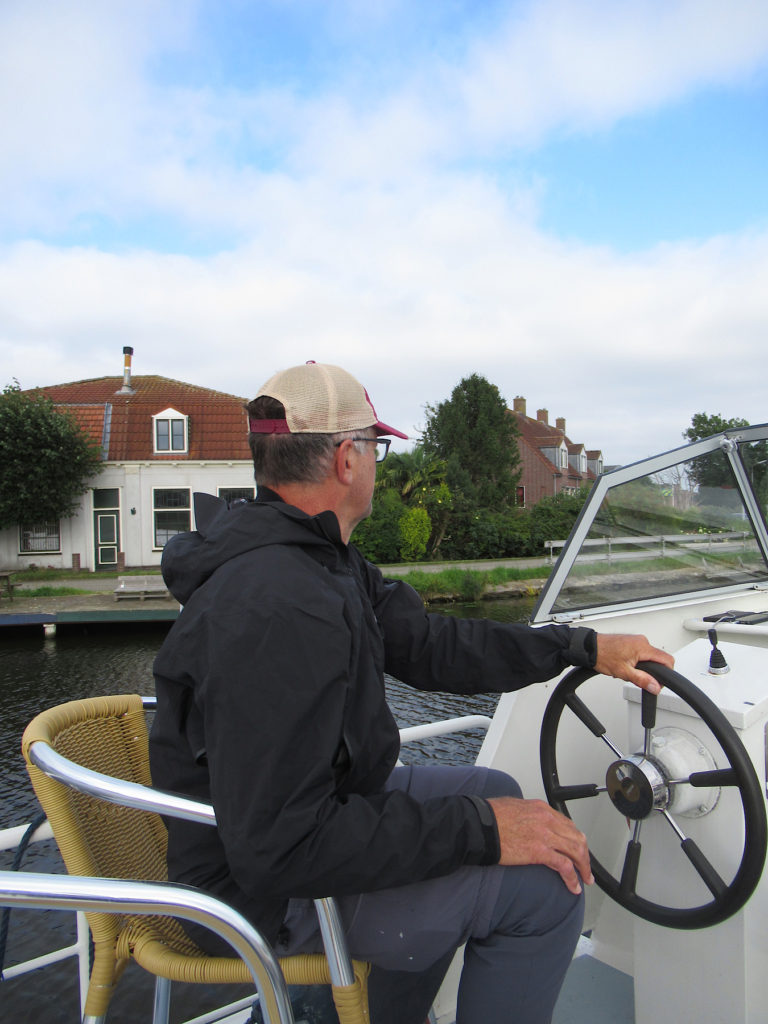 Man in black raincoat and grey pants sitting on wicker chair while steering a boat on a canal.