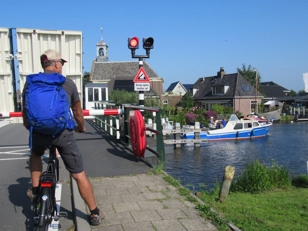 Man on bicycle wearing grey shorts with blue pack, ball cap and sunglasses waits at open bridge while a blue and white passes on canal below.