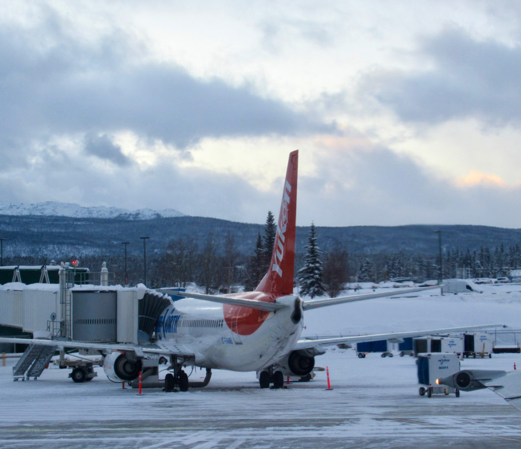 Jet with bright orangey-red pat on tail and word Yukon in white at gate in an airport.