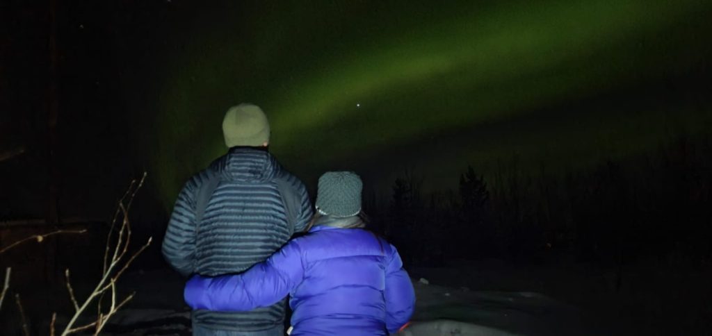 Man and woman standing in winter coats with backs to camera looking at northern lights overhead.