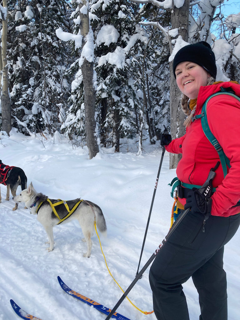 Woman in bright red jacket, dark toque and pants on skis with poles and two dog harnessed in front of her on trail.