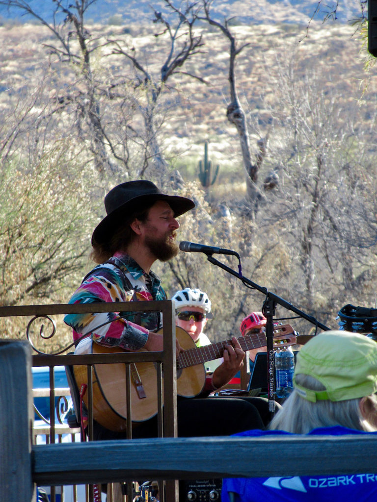 Guitar player sitting outdoors on a stage in front of a microphone wearing plaid shirt, black pants and a black cowboy hat.