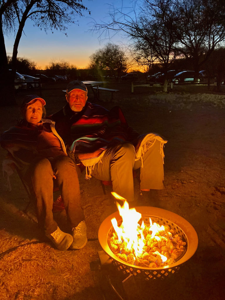 Couple sitting in lawn chairs wrapped in a colourful blanket with a propane fire burning in front of them and sunset glowing in background.