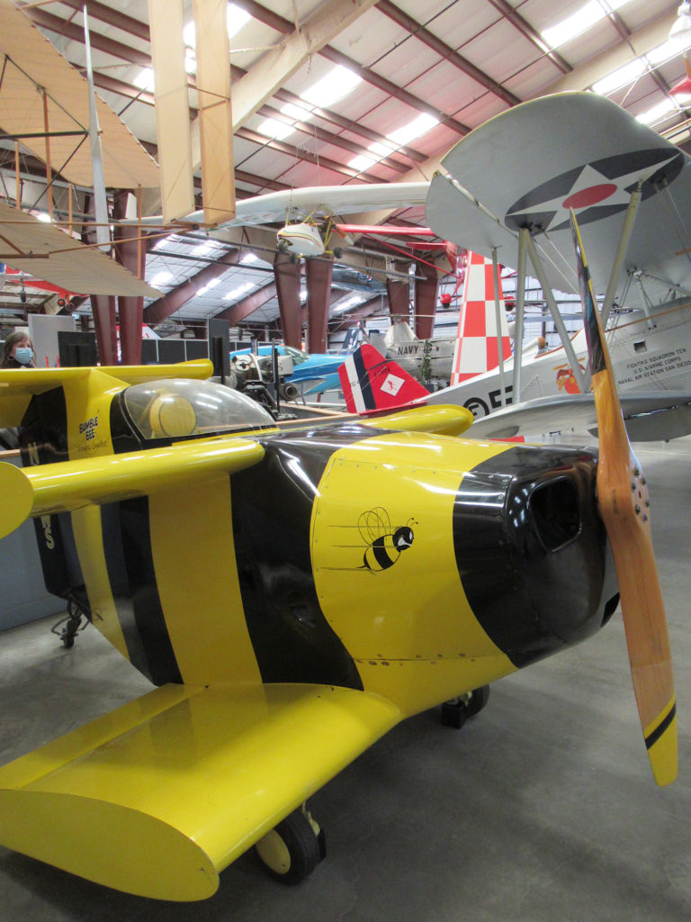 Tiny black and yellow single-person airplane with wooden propeller sitting on floor of a massive hangar filled with other aircraft in background.
