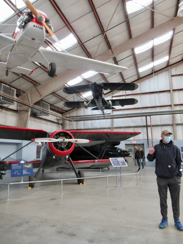 Man standing on concrete floor of massive hangar pointing up at white plane suspended from ceiling.