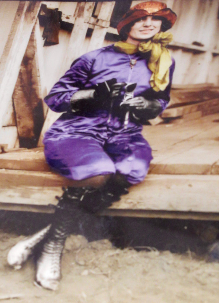 Woman sitting on wooden step wearing purple silk on piece outfit with high black boots, yellow scarf and orange hat.