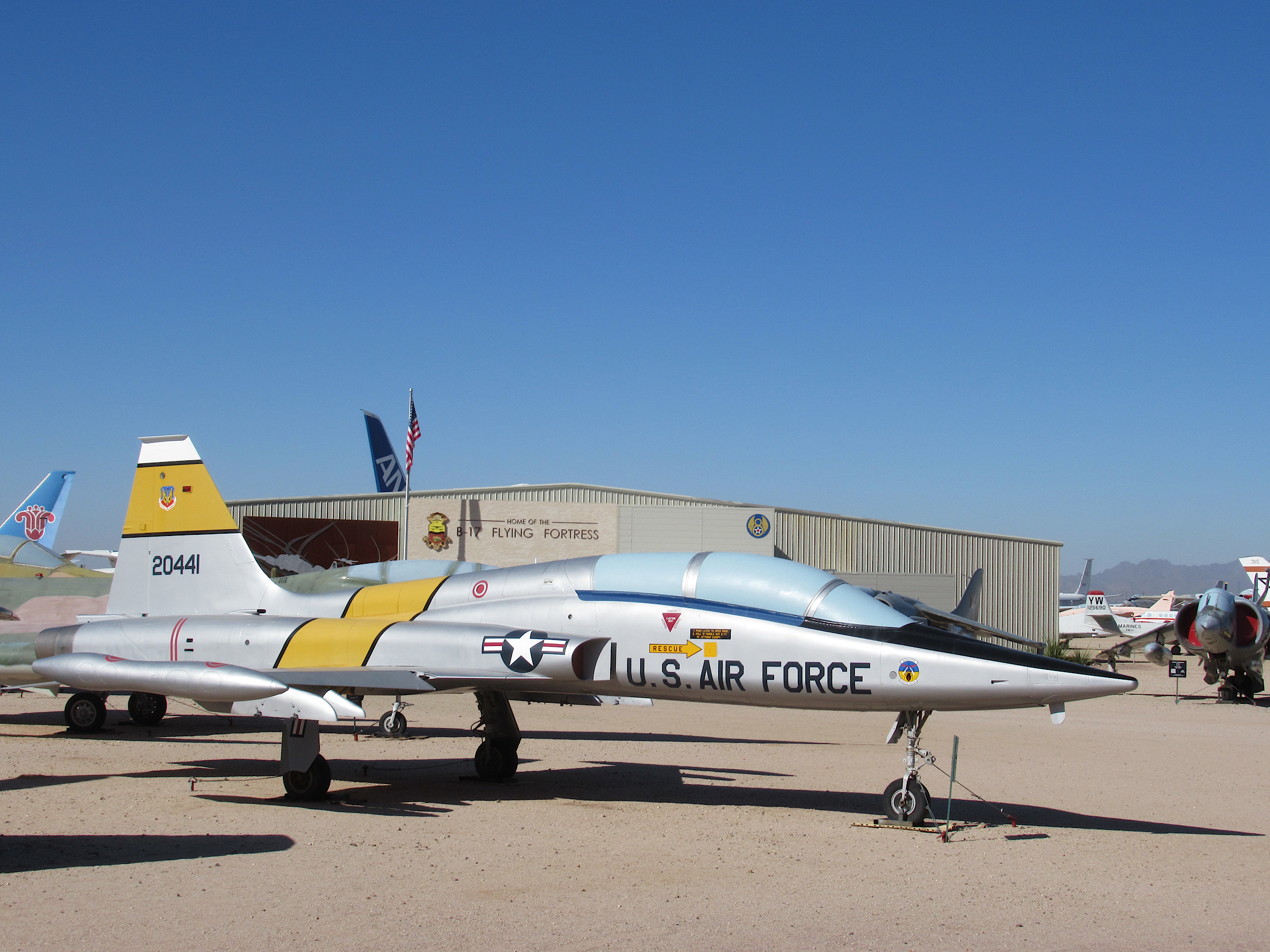 U.S. Air Force fighter plane with yellow and black paint on gravel in front of large hangar.