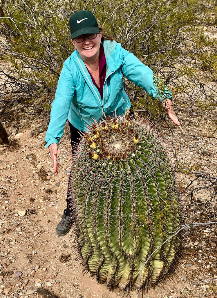 Woman in light blue windbreaker and black ball cap pretending to hug a barrel cactus.