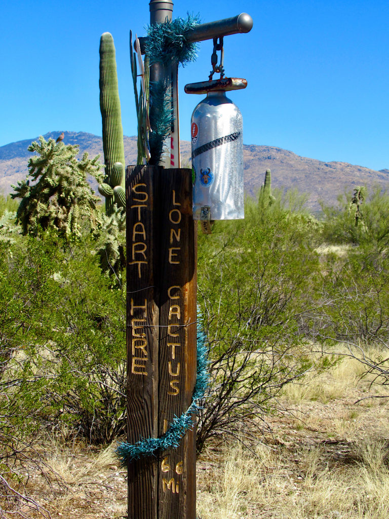 Silver bell hanging from a wooden post engraved with the words: Start here Lone Cactus 6.6 miles