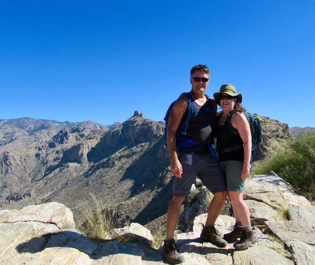 Man and woman in hiking shorts and short sleeved tops standing on rocky ridge with mountains in background above their RV Park campsite.
