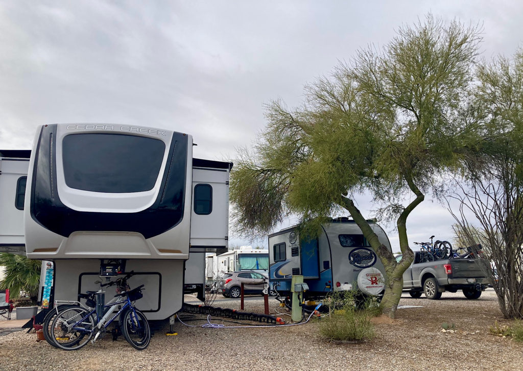 Large fifth wheel with many slideouts next to smaller blue Rpod trailier parked behind a greenish Palo Verde tree.